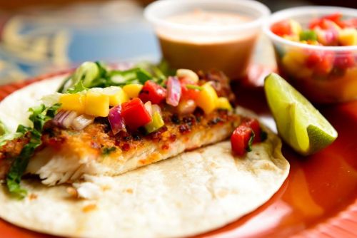 A fish taco topped with colorful salsa, served with lime and two small cups of sauce and salsa on a vibrant plate.