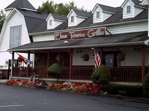 Exterior of Elma Towne Grille featuring a porch, flower beds, and flags, set against a cloudy sky.