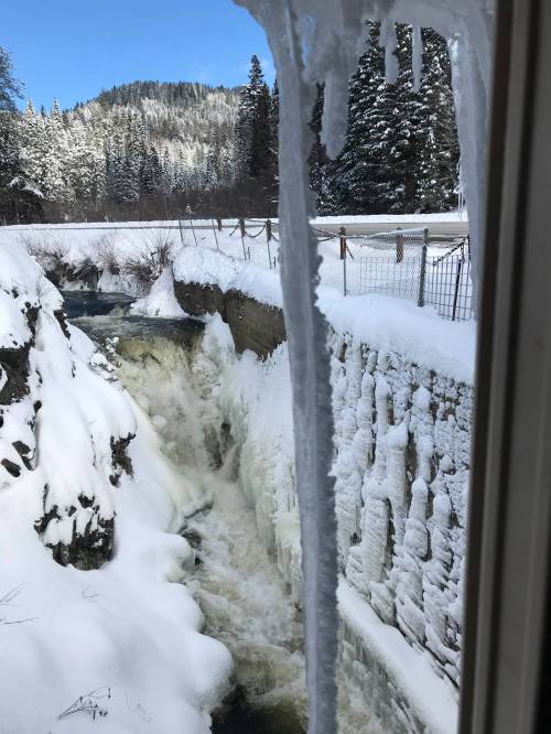 Icicles hang from a window, overlooking a snowy landscape and a rushing waterfall surrounded by trees.