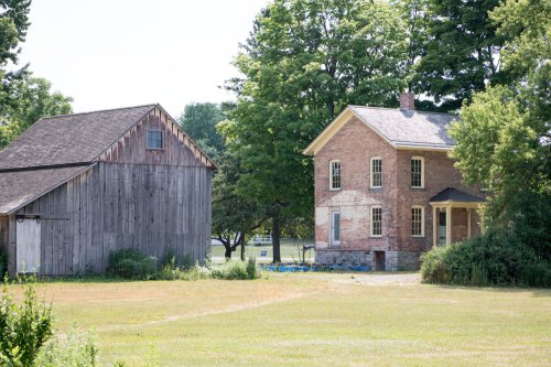 A rustic wooden barn and a brick house surrounded by green trees and grass on a sunny day.