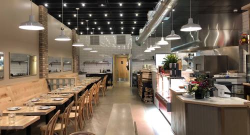 Interior of a modern restaurant featuring wooden tables, bright lighting, and an open kitchen area.