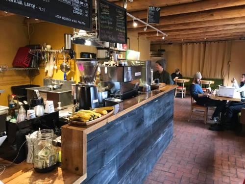 A cozy café interior with a wooden counter, coffee equipment, and patrons seated at tables. Bananas are displayed on the counter.