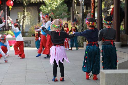 Children in colorful costumes dance joyfully in a festive outdoor setting, surrounded by decorations and other performers.
