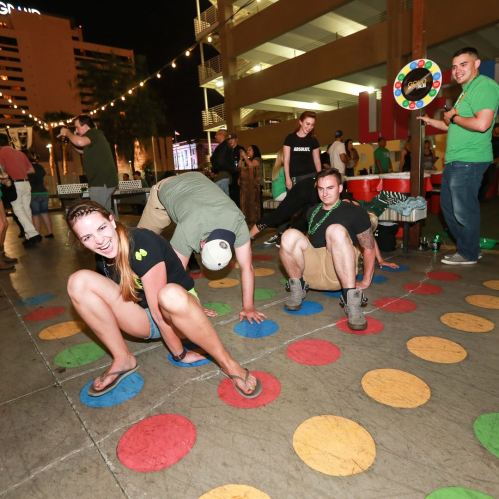 A group of people playing Twister outdoors at night, with colorful circles on the ground and festive lights in the background.