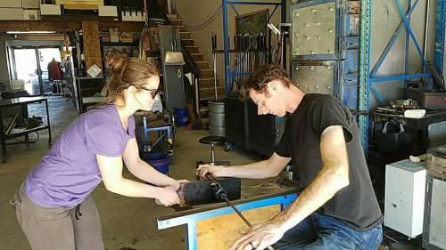 Two people work together in a workshop, shaping metal with tools at a workbench.