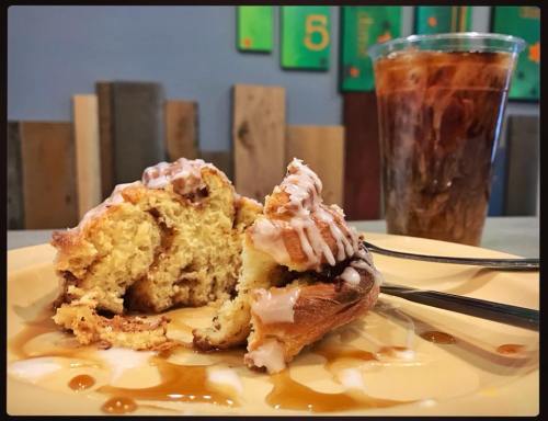 A plate with a cinnamon roll drizzled with icing, alongside a glass of iced coffee on a wooden table.