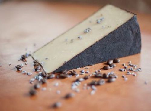 A wedge of cheese with a dark rind, sprinkled with coffee beans and salt, resting on a wooden surface.