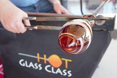 A person shaping a colorful glass orb using glassblowing tools at a workshop.