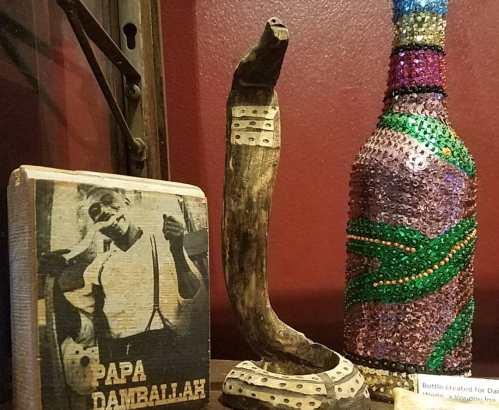A wooden snake sculpture, a decorative bottle, and a book titled "Papa Damballah" displayed on a shelf.