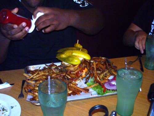 A person adds ketchup to a large burger topped with pickles, surrounded by a mountain of fries and drinks on the table.