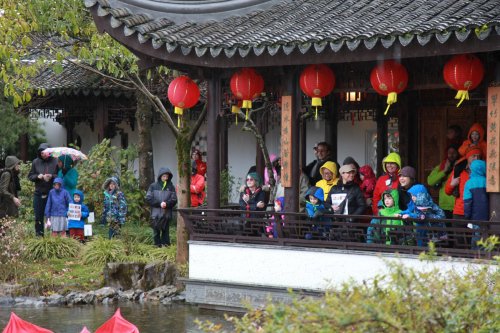 A crowd of people in colorful raincoats stands by a pond, enjoying a festive event with red lanterns overhead.