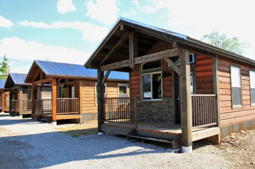 A row of rustic wooden cabins with porches, set against a blue sky and surrounded by gravel pathways.