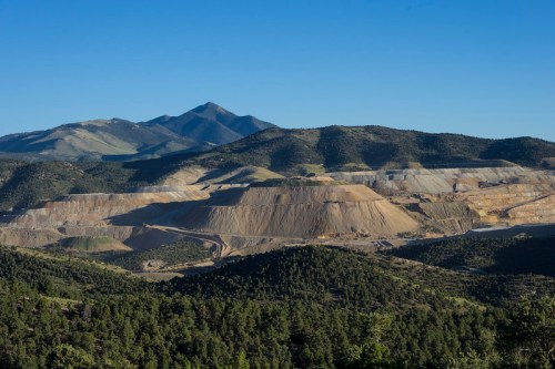 A vast mining site surrounded by green forests and mountains under a clear blue sky.