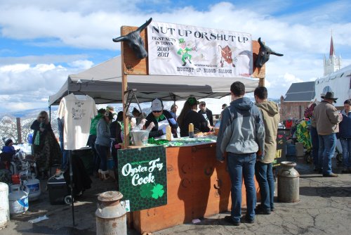 A busy outdoor festival booth with people enjoying food and drinks, decorated with festive banners and St. Patrick's Day themes.