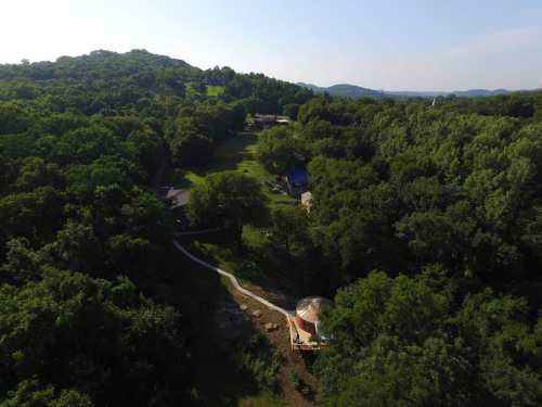 Aerial view of a lush green landscape with a winding path and a yurt surrounded by trees and hills in the background.