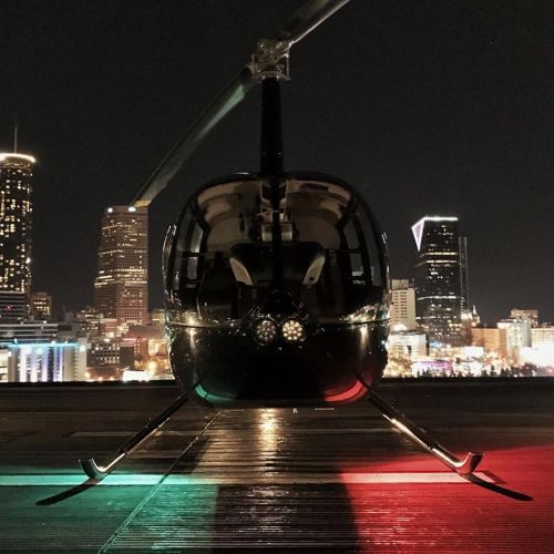 A black helicopter on a rooftop at night, with city skyline lights and red and green lights on the landing skids.