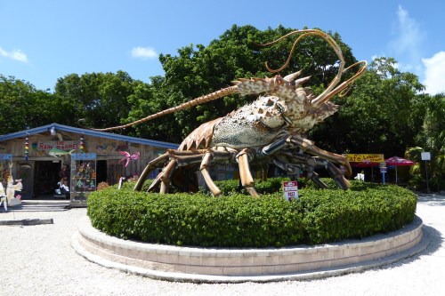 A large, colorful lobster sculpture surrounded by greenery, with a building and blue sky in the background.