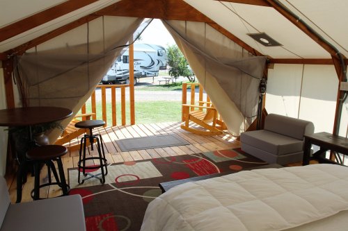 Interior of a glamping tent with a bed, seating area, and an open entrance leading to a wooden deck.