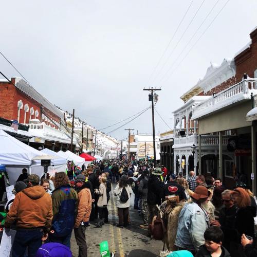 A bustling street festival with crowds, tents, and historic buildings under a cloudy sky. Snow-capped rooftops in the background.