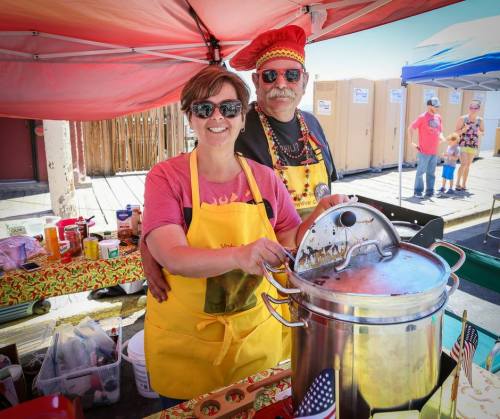 A smiling woman in a yellow apron and a man in a red hat stand behind a cooking pot at a festive outdoor market.