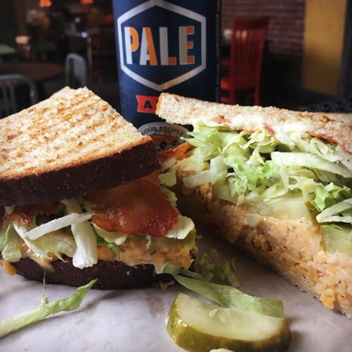 A close-up of a sandwich with lettuce, bacon, and a creamy filling, served with pickles and a can of pale ale.