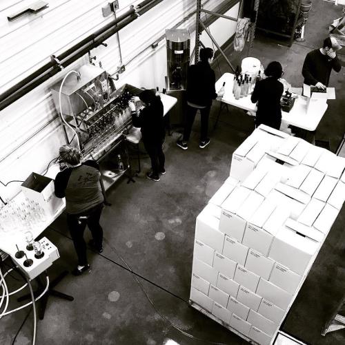 Workers in a warehouse bottling and packaging products, with stacked boxes in the foreground and equipment in the background.