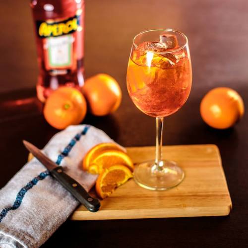 A glass of orange cocktail with ice, surrounded by oranges and a knife on a wooden cutting board.