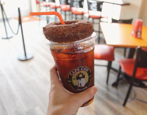A hand holding a cup of iced coffee topped with a cinnamon sugar donut, with a café background.