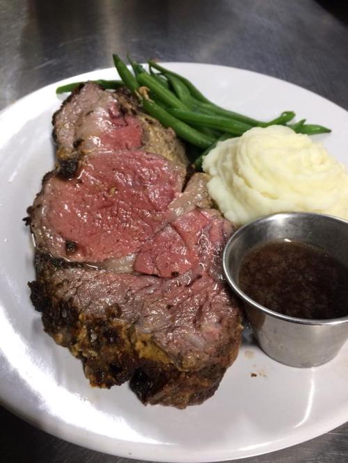 A plate with a slice of medium-rare roast beef, mashed potatoes, green beans, and a small cup of gravy.
