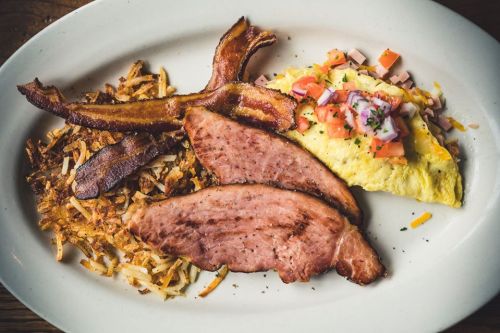 A plate of breakfast featuring bacon, ham slices, an omelet with tomatoes, and crispy hash browns.