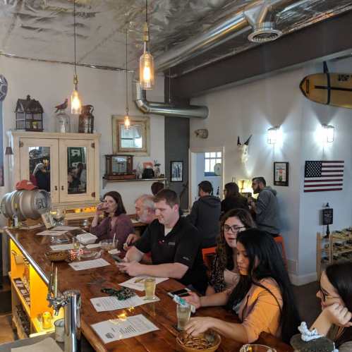 A lively bar scene with patrons enjoying drinks and food at a wooden counter, surrounded by decor and warm lighting.