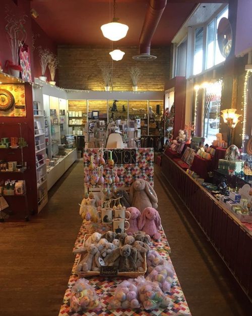 A colorful gift shop aisle filled with plush toys, cards, and decorative items, featuring bright lighting and wooden floors.