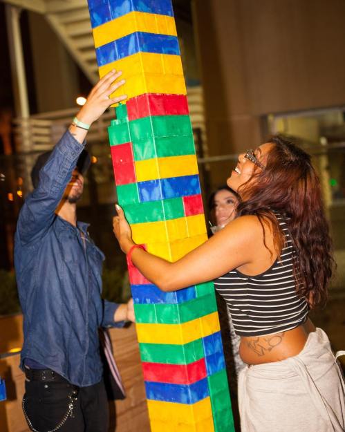 Two people are stacking colorful blocks while a third person smiles in the background, enjoying the playful activity.