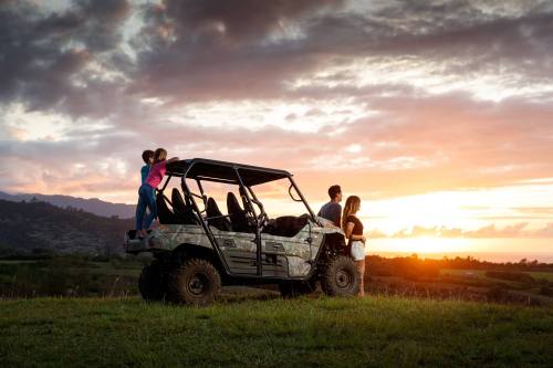 A family enjoys a sunset view beside a camouflaged off-road vehicle on a grassy hill.
