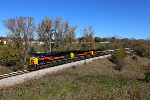A freight train with black and yellow engines travels along a tree-lined track under a clear blue sky.