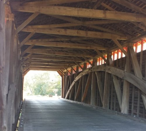 Interior view of a wooden covered bridge, showcasing its beams and structure, with light streaming in from the entrance.