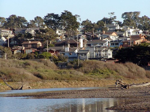 A scenic view of houses on a hillside, surrounded by trees and a calm river in the foreground.