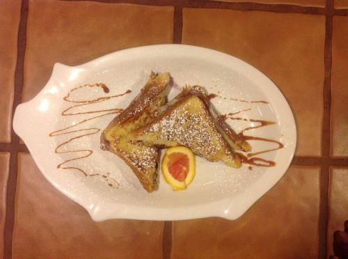 A plate of French toast triangles dusted with powdered sugar, drizzled with sauce, and garnished with an orange slice.