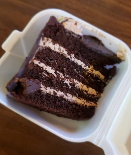 A slice of chocolate cake with layers of frosting, placed in a white takeout container.