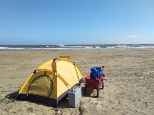 A yellow tent and a red cart on a sandy beach with ocean waves and a clear blue sky in the background.
