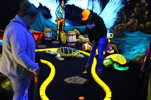 Two people playing mini-golf in a glow-in-the-dark setting with colorful decorations and sea-themed elements.