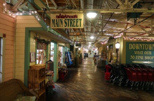 A charming indoor market with shops lining a wooden walkway, decorated with lights and a "Main Street" sign.