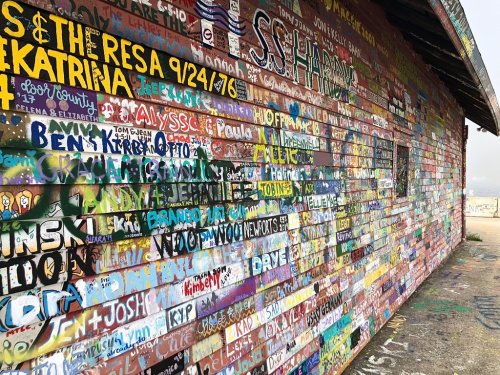 Colorful graffiti covers a brick wall, featuring names, dates, and messages in various styles and colors.