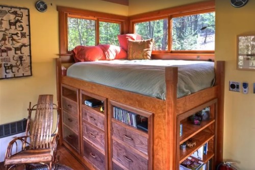 Cozy loft bed with orange pillows, surrounded by windows, and wooden storage drawers beneath in a bright room.
