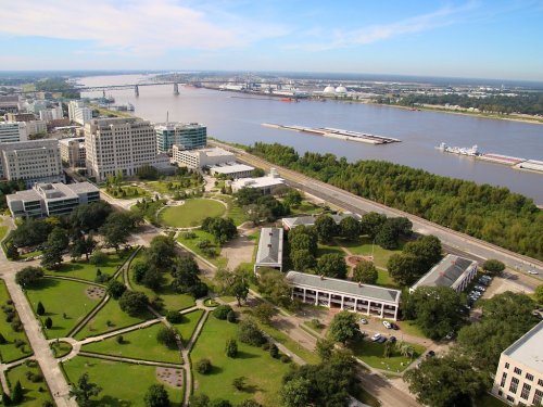 Aerial view of a cityscape with green parks, a river, and industrial areas in the background under a clear blue sky.