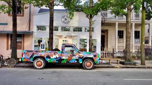 A colorful, camouflaged truck parked in front of a quaint building with large windows and trees lining the street.