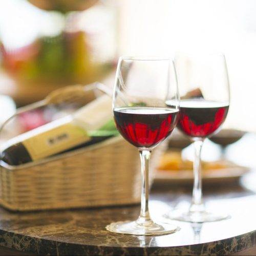 Two glasses of red wine on a marble table, with a basket of wine bottles in the background.