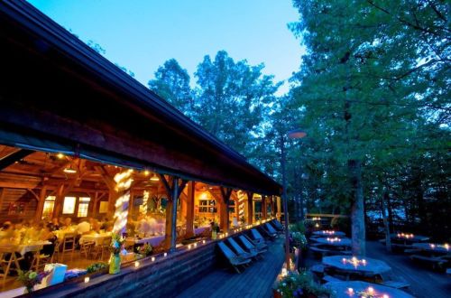 A cozy outdoor dining area at dusk, featuring a wooden cabin, tables, and candles surrounded by trees.