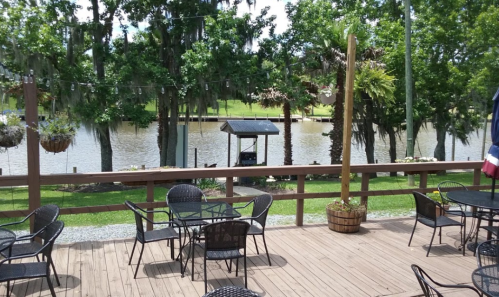 A wooden deck with empty tables and chairs overlooks a calm river surrounded by trees and greenery.