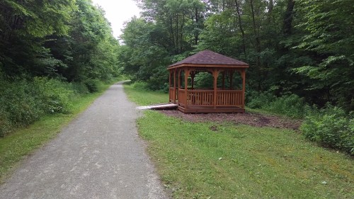 A wooden gazebo beside a gravel path, surrounded by lush green trees and foliage.